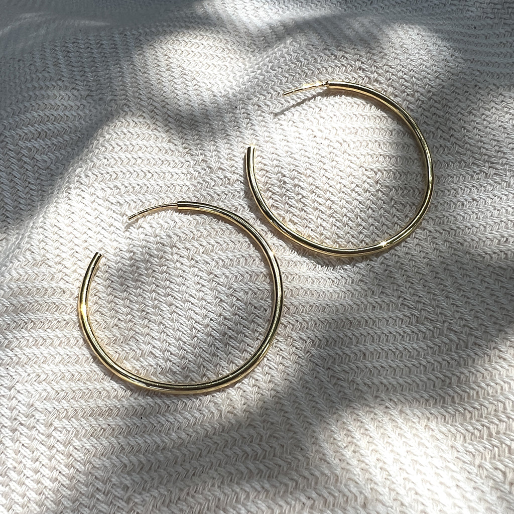 Pair of large gold plated hoop earrings on woven linen under leaf shadows