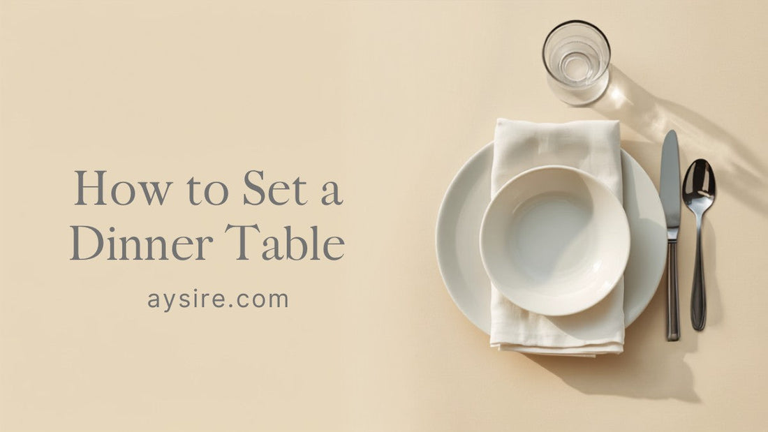 Overhead flat lay of a single place setting on a warm beige table: white porcelain plate and bowl, stainless steel knife and spoon to the right, a folded linen napkin and a glass above