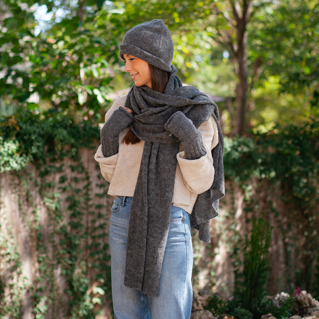 Woman outdoors in a charcoal beanie, gloves and a long charcoal knit scarf, cream sweater and blue jeans