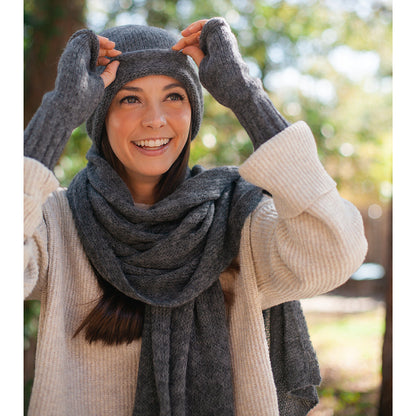 Smiling woman pulling on a charcoal beanie with both hands, charcoal fingerless gloves and scarf on
