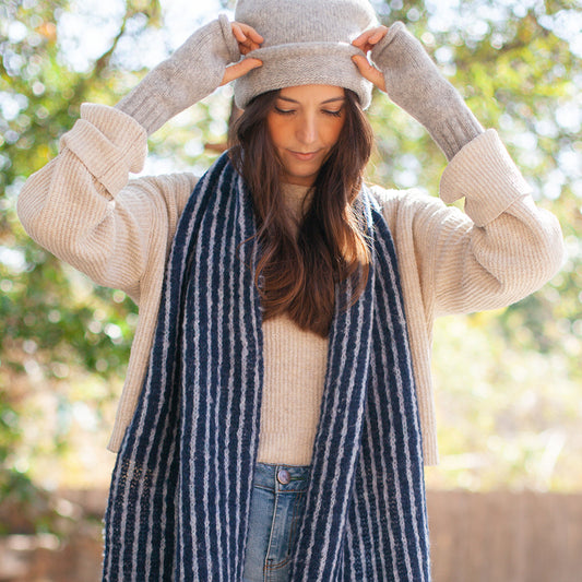 Woman in a light gray beanie, gray wrist warmers and a navy and white striped scarf, hands raised to the hat
