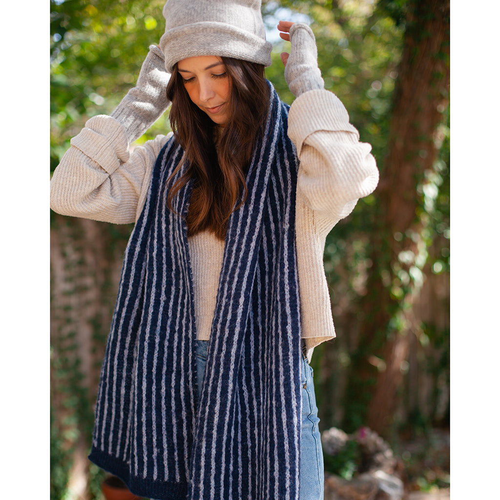 Woman in a light gray beanie and gray wrist warmers, hands on the hat, a navy and white striped scarf around her neck