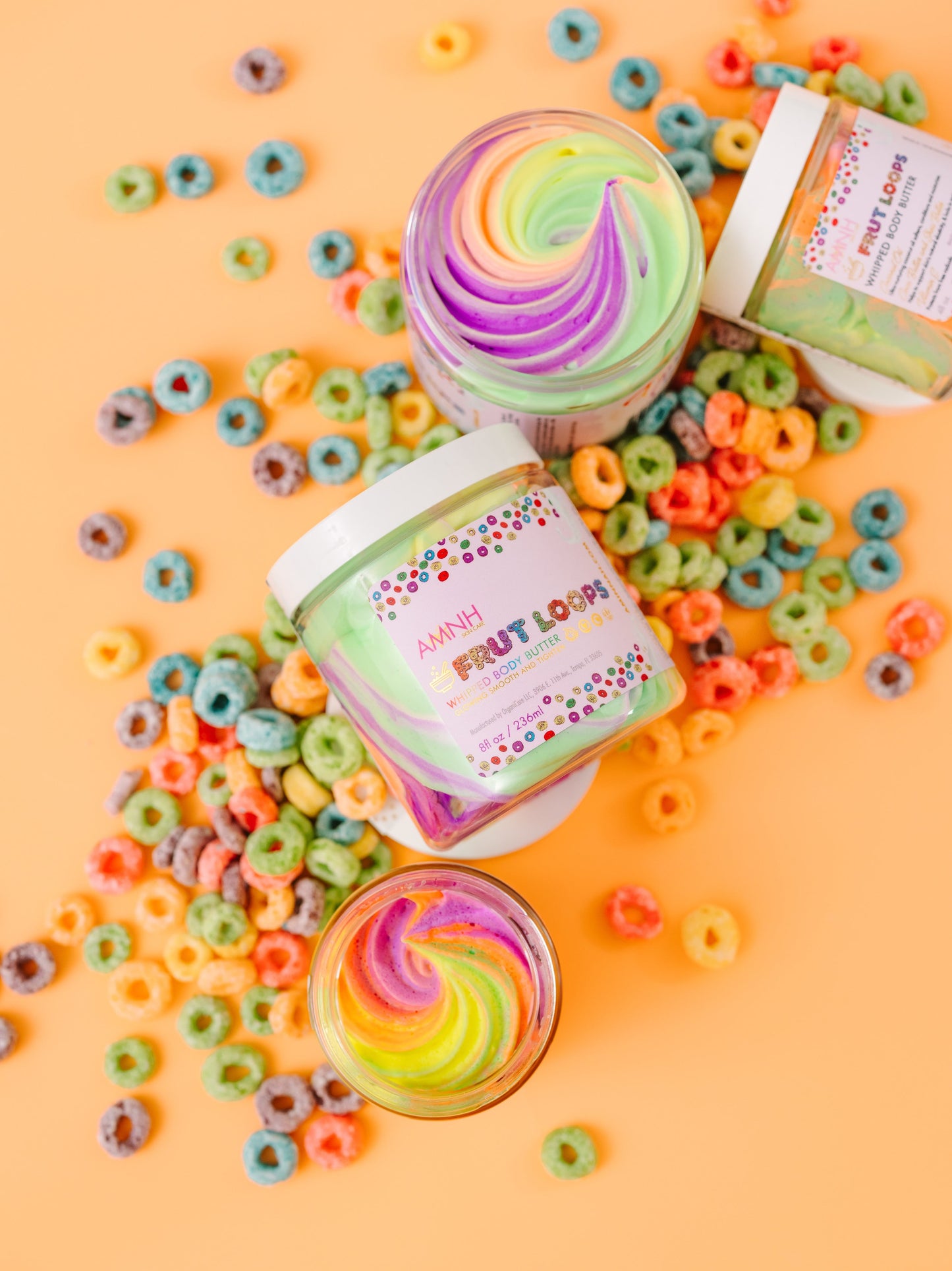 Three open jars of rainbow whipped butter among cereal loops and sprinkles on orange.
