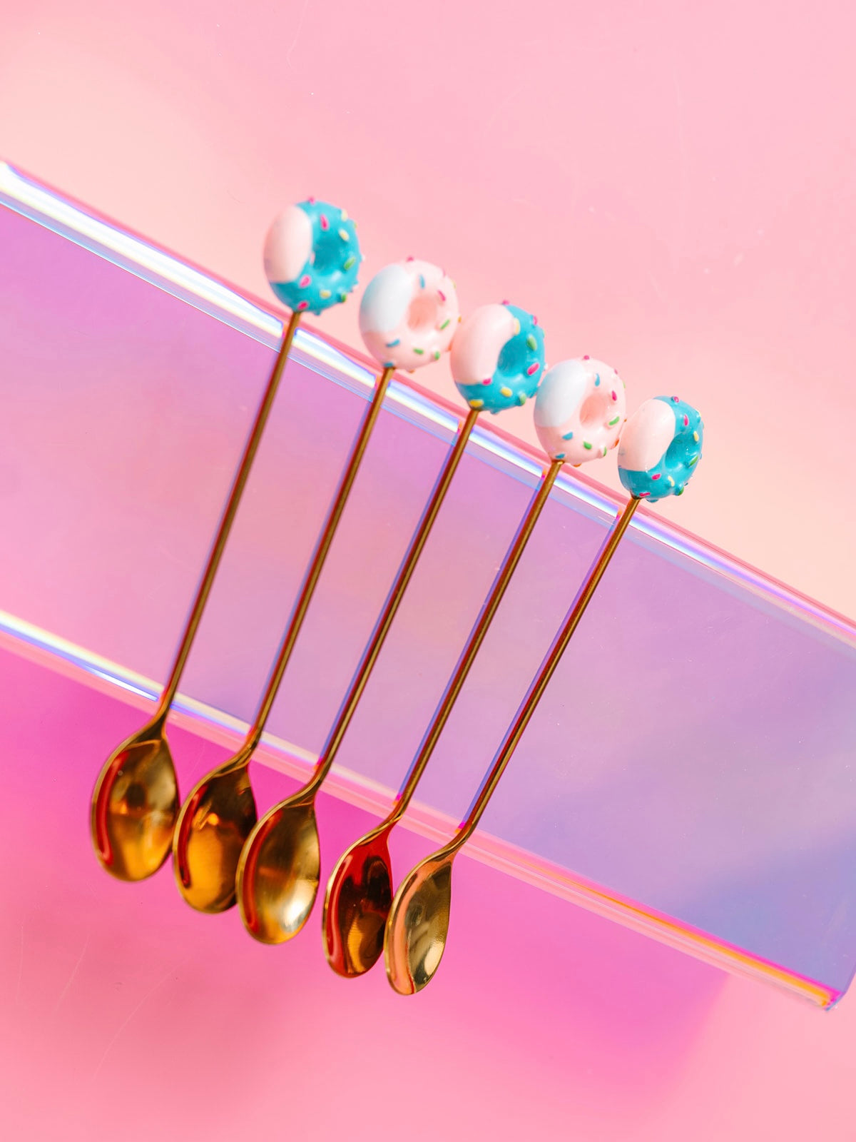 Five gold spoons with donut toppers laid in a row on an iridescent surface.