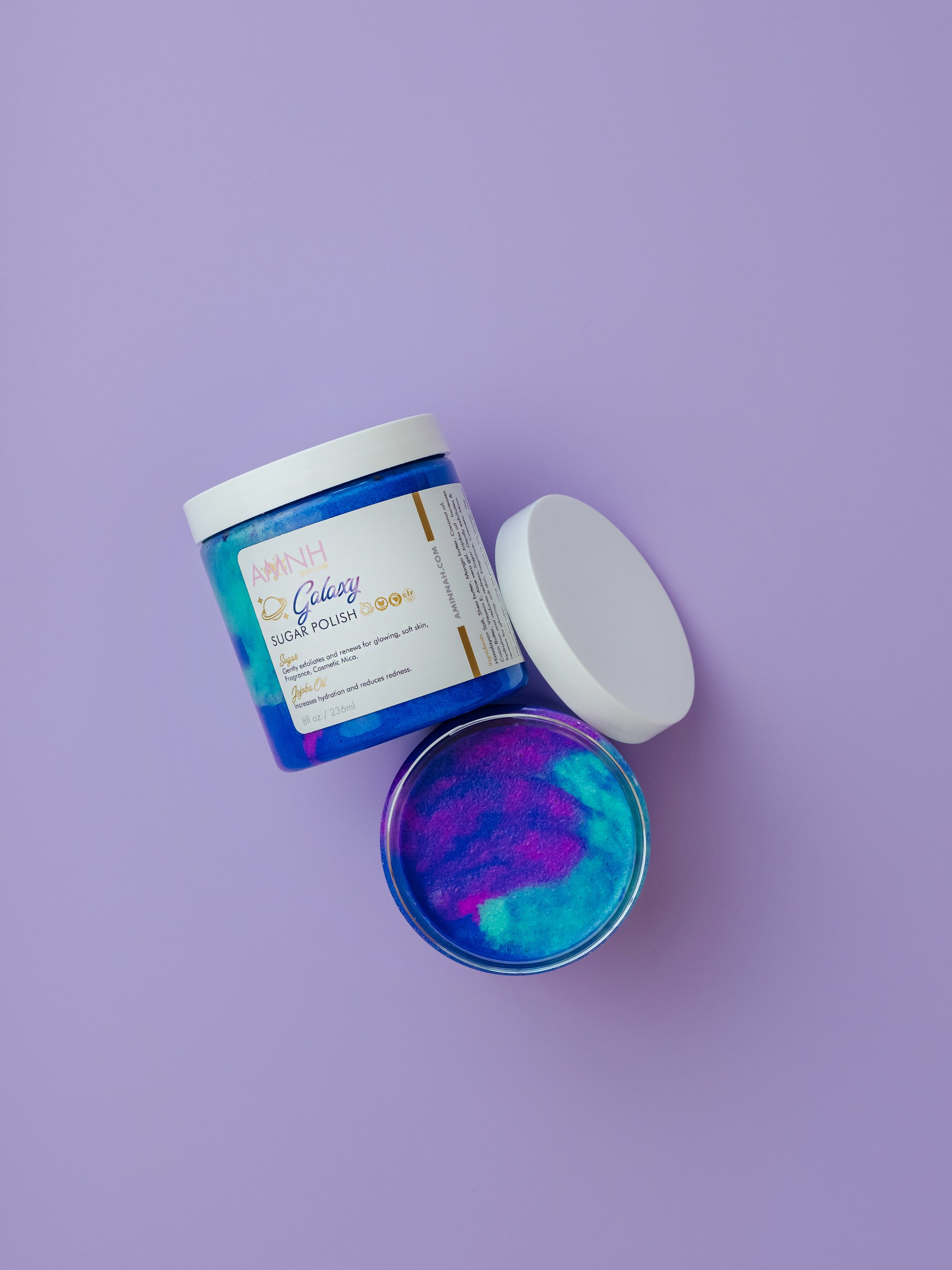 Jar of galaxy scrub with the lid propped beside it on purple.