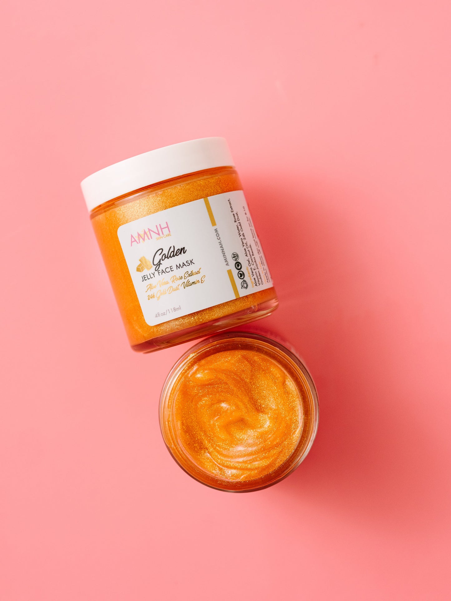 Open jar of gold jelly face mask with the lid resting beside it on a pink background.