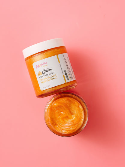 Open jar of gold jelly face mask with the lid resting beside it on a pink background.