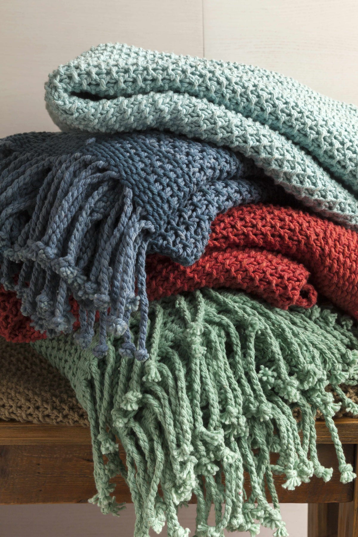 Knit throws in aqua, blue and red piled on a bench with twisted fringe hanging over the edge