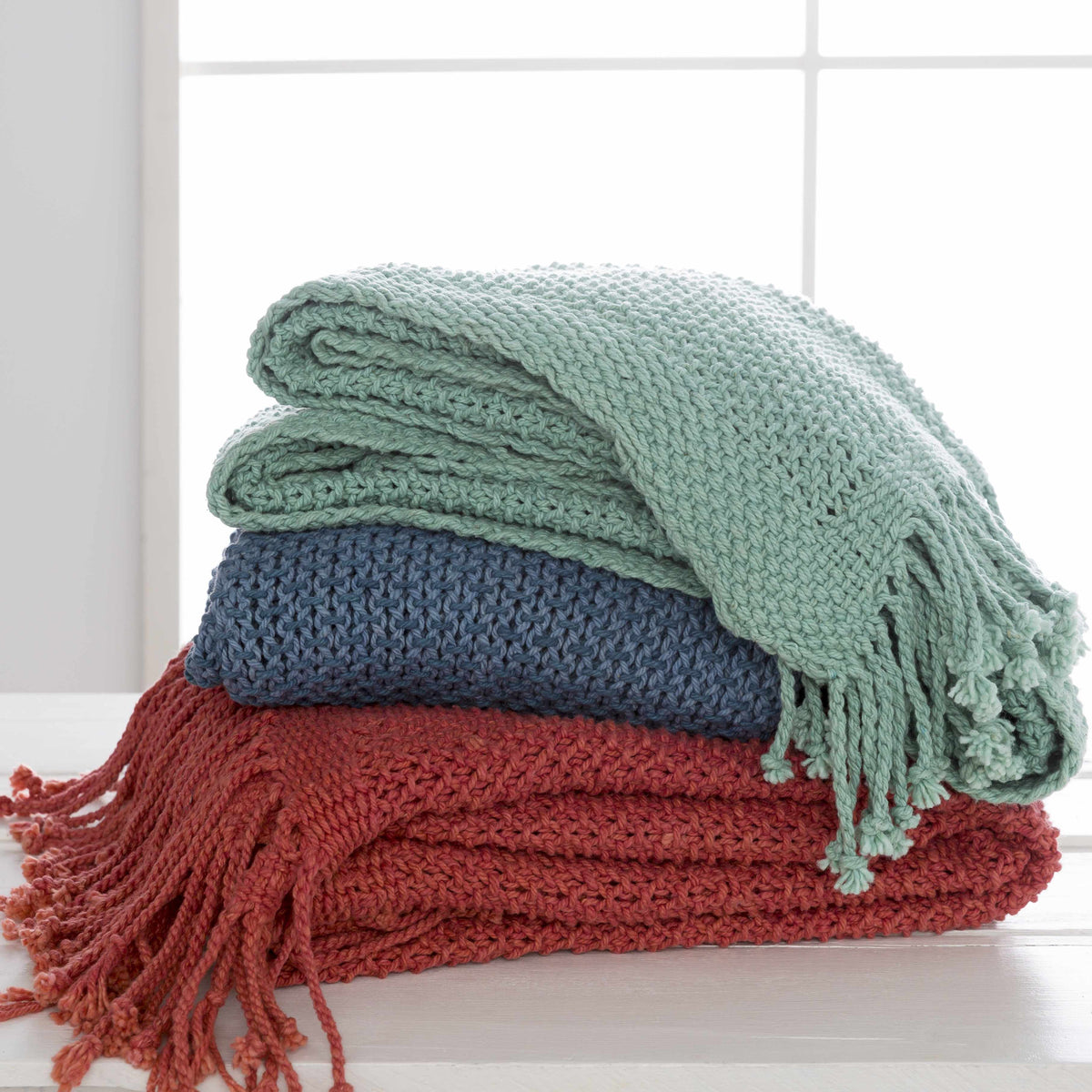 Aqua green knit throw folded on top of blue and red knit throws in front of a window