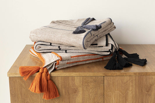 Three folded knit throws stacked on a wood cabinet, with black, gray and orange corner tassels