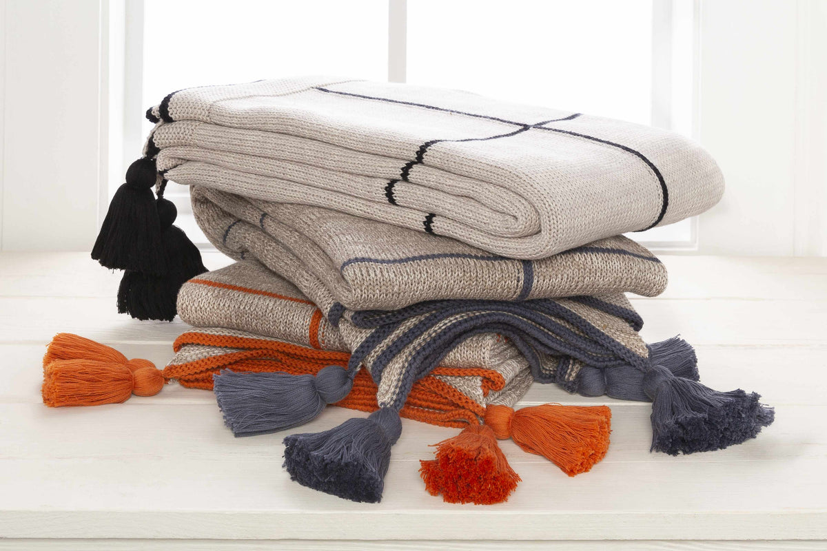 Stack of folded cream knit throws with black, gray and orange tassels on a white table