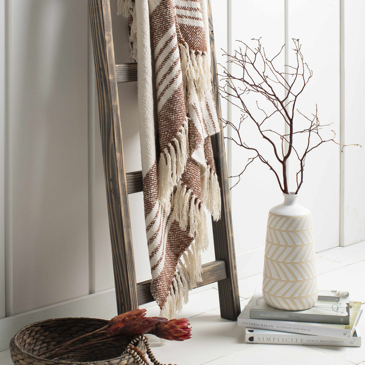 Cream and rust striped throw blanket draped on a wooden ladder beside a white vase with bare branches