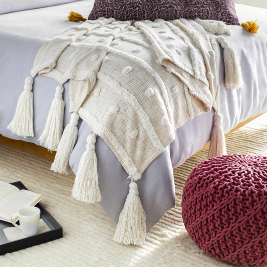 Cream tufted cotton throw blanket with long tassels draped over the end of a bed beside a knit pouf
