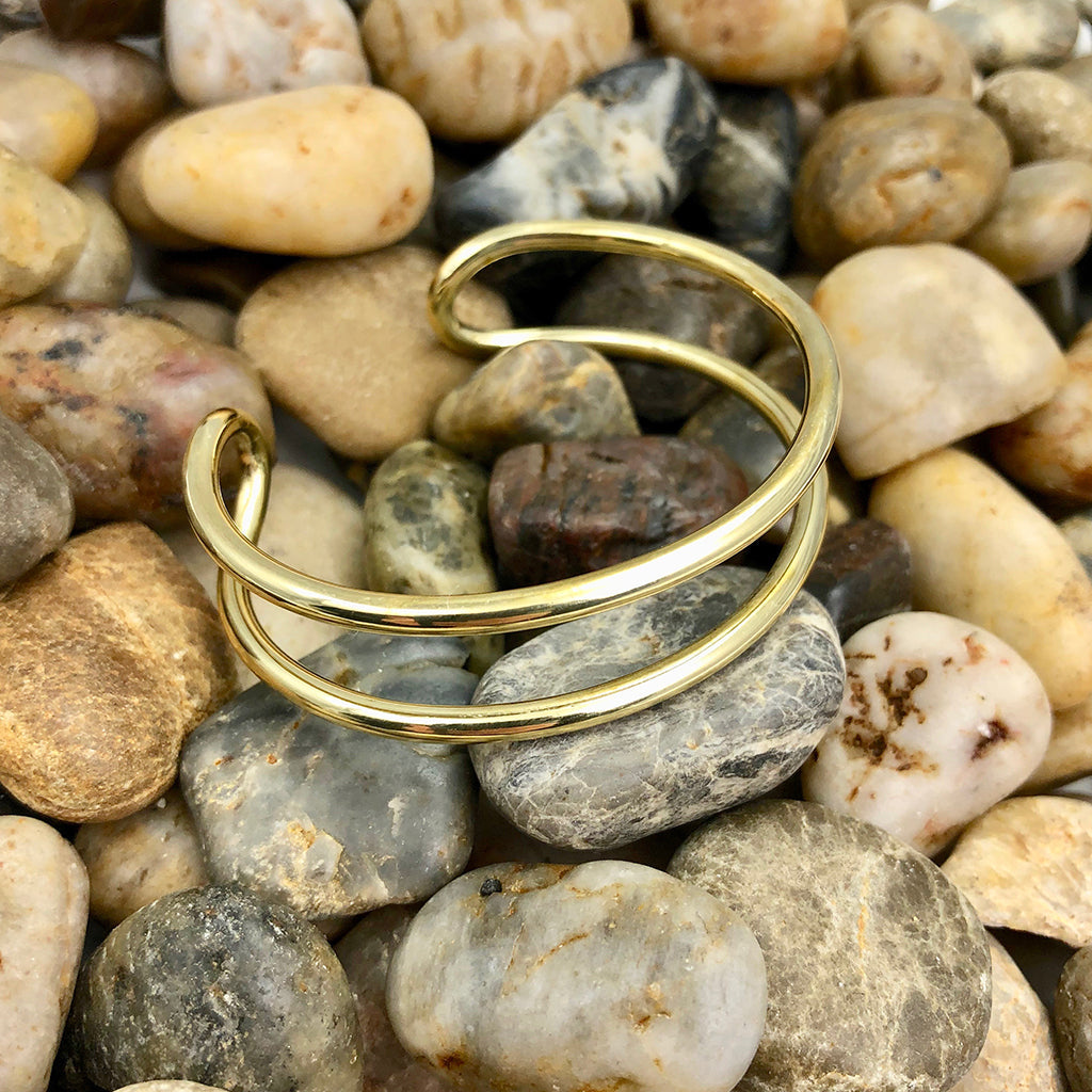 Double wire brass cuff bracelet resting on smooth river pebbles in brown and gray