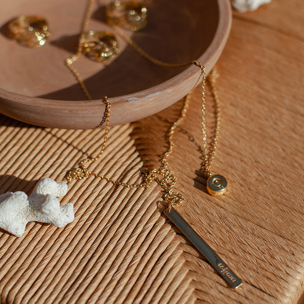 Layered bar necklace on a woven rush seat beside a clay bowl and white coral