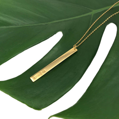 Engraved brass bar of the layered necklace lying on a green monstera leaf