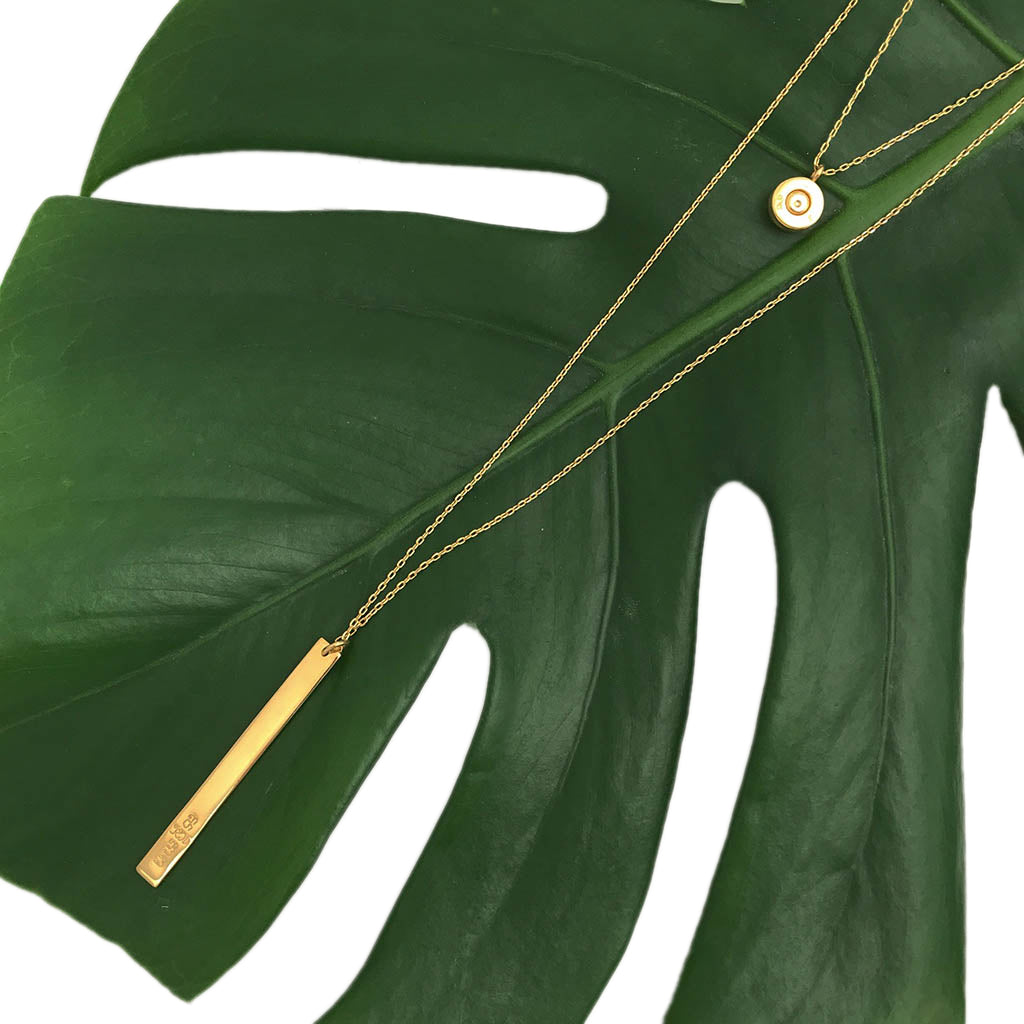 Layered bar necklace across a monstera leaf, the round charm above the long bar
