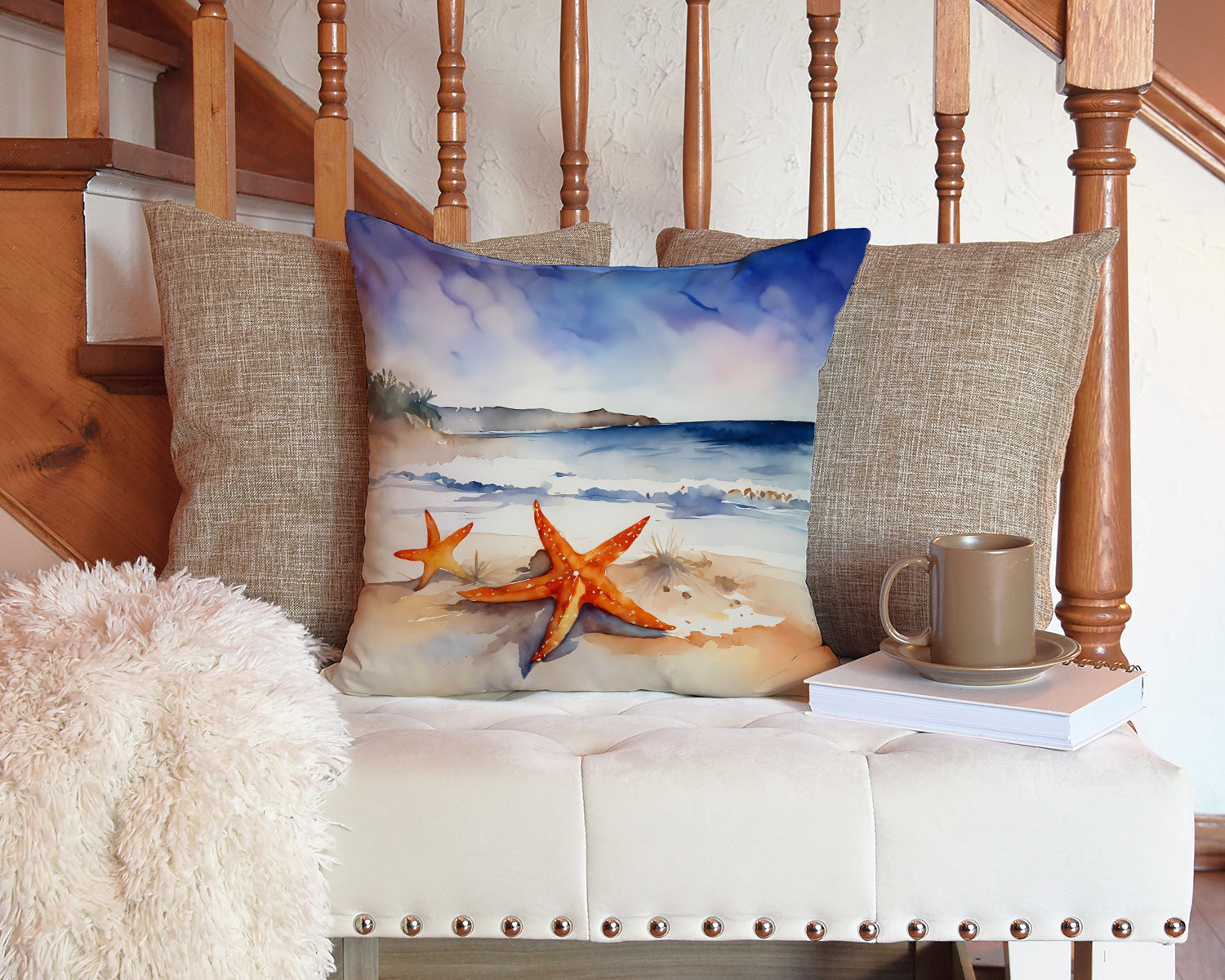 The Two starfish artwork pillow on a white indoor bench at the foot of a staircase, beside a throw and a mug.