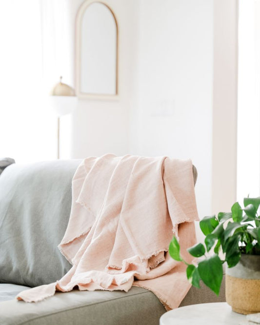 Blush linen throw blanket draped over the arm of a gray sofa beside a potted plant