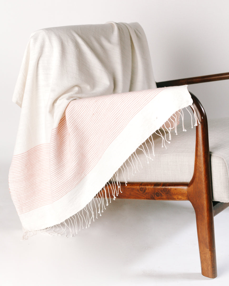 Natural cotton throw with blush stripes draped over a wood frame armchair, fringe hanging toward the floor