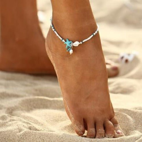 White beaded anklet with a turquoise starfish and shell charm worn on a bare foot in the sand