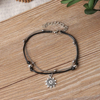 Sun anklet laid on a wooden table with its black cord, silver beads, chain, sun charm and extender