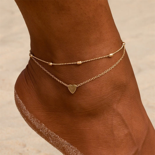 Gold tone double chain heart anklet worn on a tanned ankle against the sand