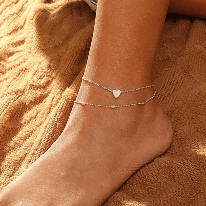 Silver tone heart anklet with two fine chains worn on a bare foot resting on a knit blanket