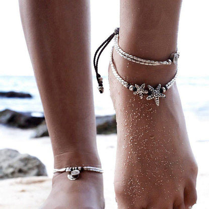 Starfish anklet with two silver bead strands worn on a sandy ankle alongside other anklets at the beach