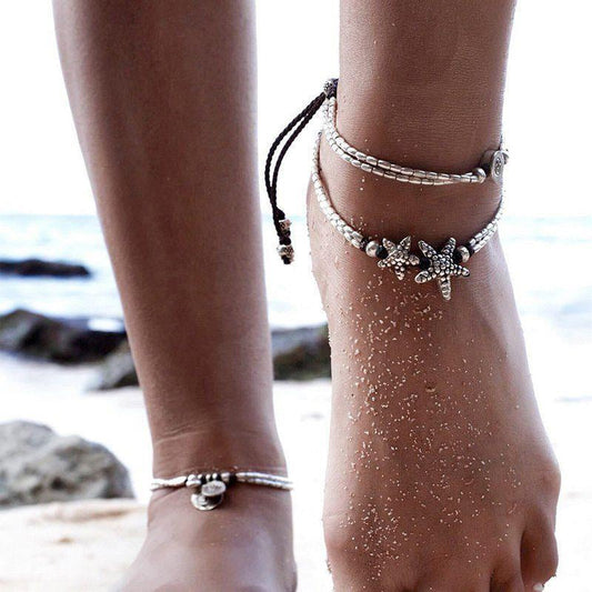 Starfish anklet with two silver bead strands worn on a sandy ankle alongside other anklets at the beach