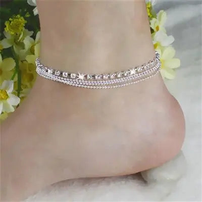 Close view of the crystal row and three chains of the layered anklet on the ankle, flowers behind