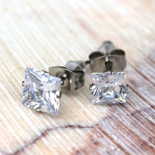 Pair of princess cut clear CZ stud earrings with four corner prongs on surgical steel posts, on wood