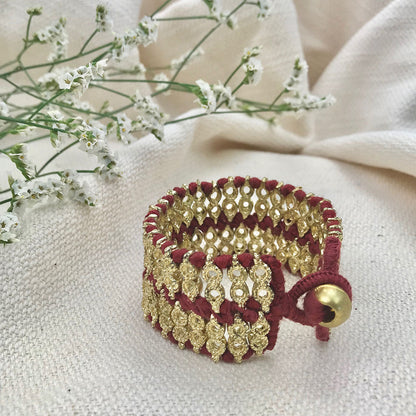 Wide woven cuff with wine red thread and gold tone metal beads beside white flowers