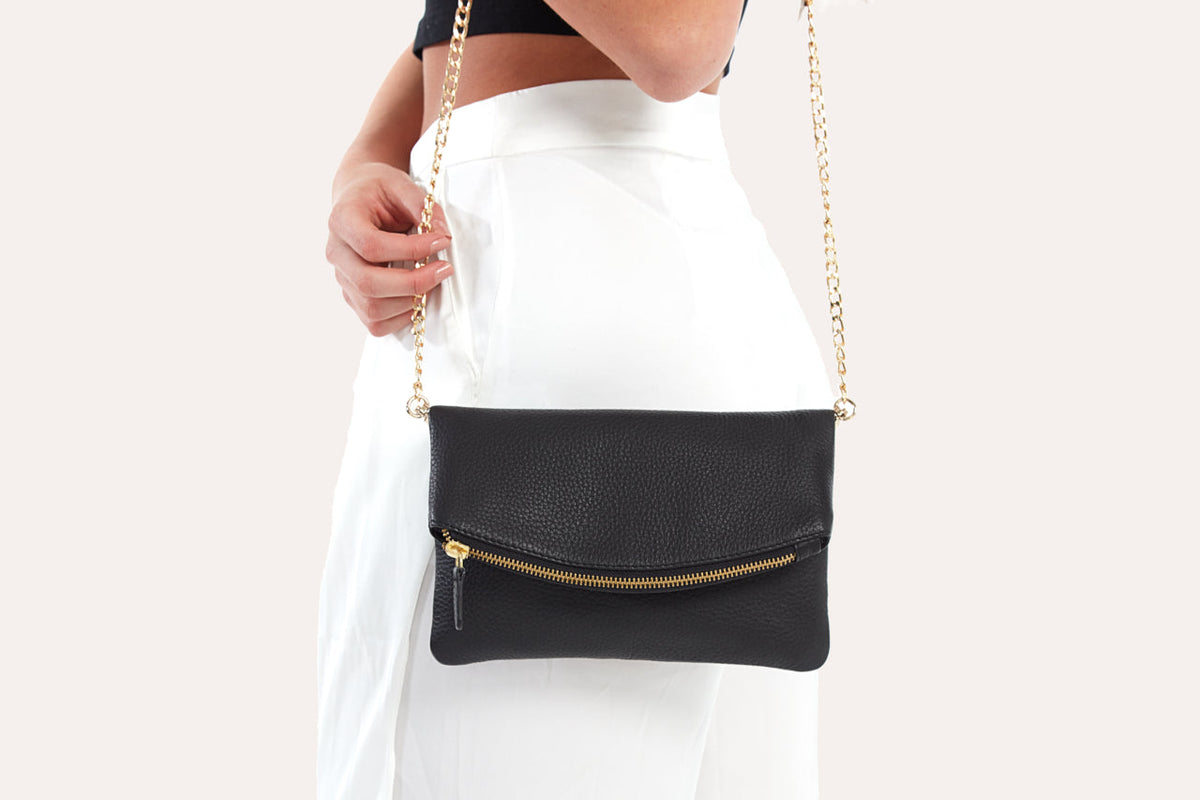 Black fold over clutch worn on the shoulder on its gold-tone chain strap against white pants
