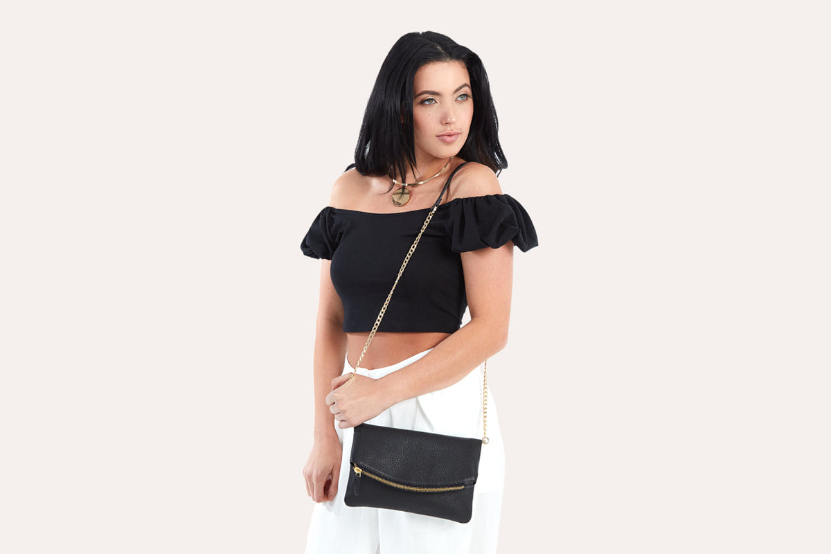 Woman in a black top and white pants with the black fold over clutch on its chain at the hip