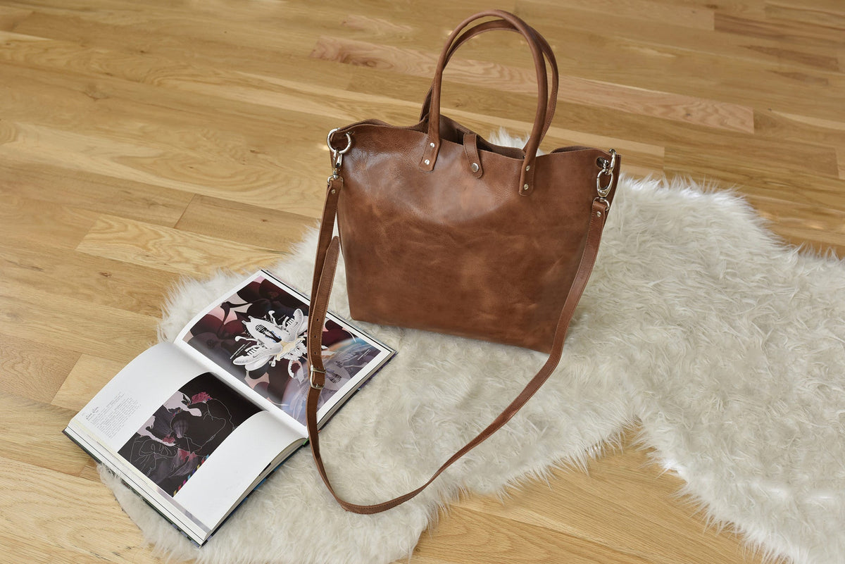 Caramel leather tote on a faux fur rug with its long shoulder strap attached, beside an open magazine