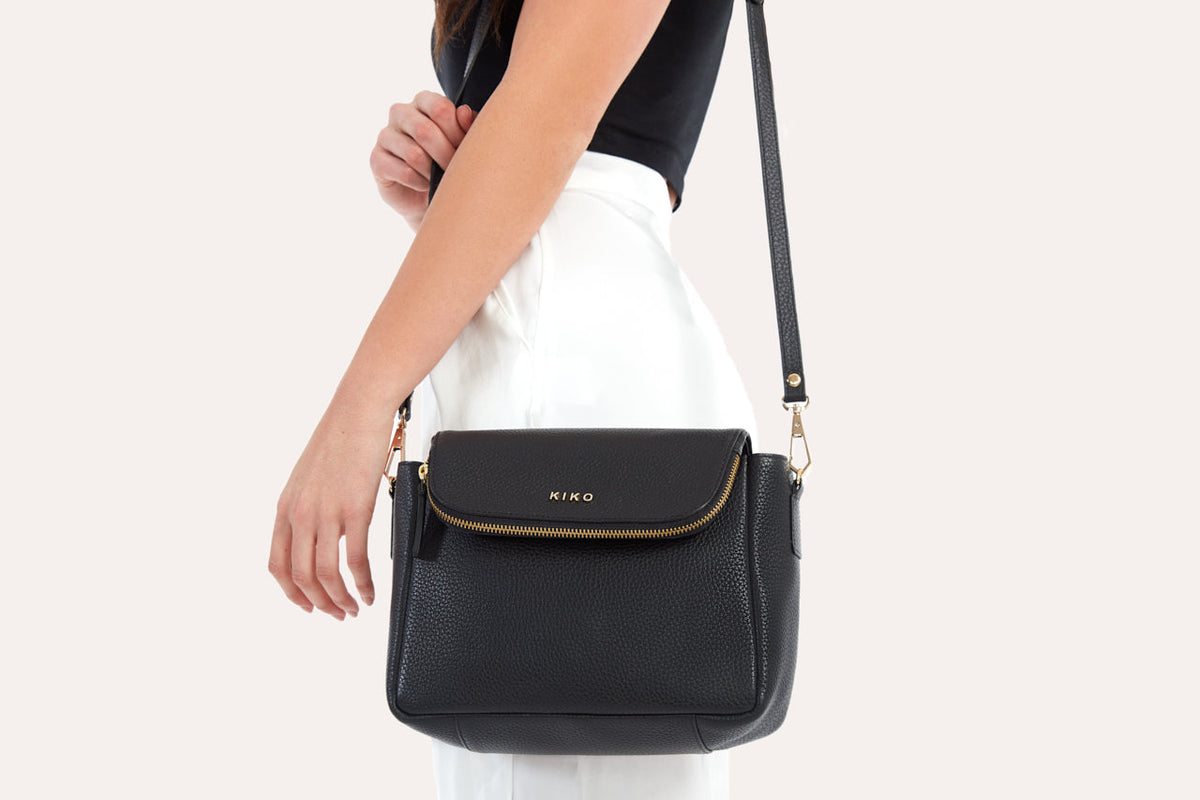 Black flap crossbody bag worn on its long adjustable strap against white pants, side view