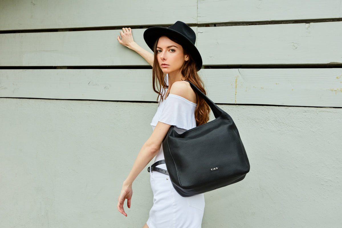 Woman in an off-shoulder top and black hat wearing the black hobo bag on her shoulder by a wall