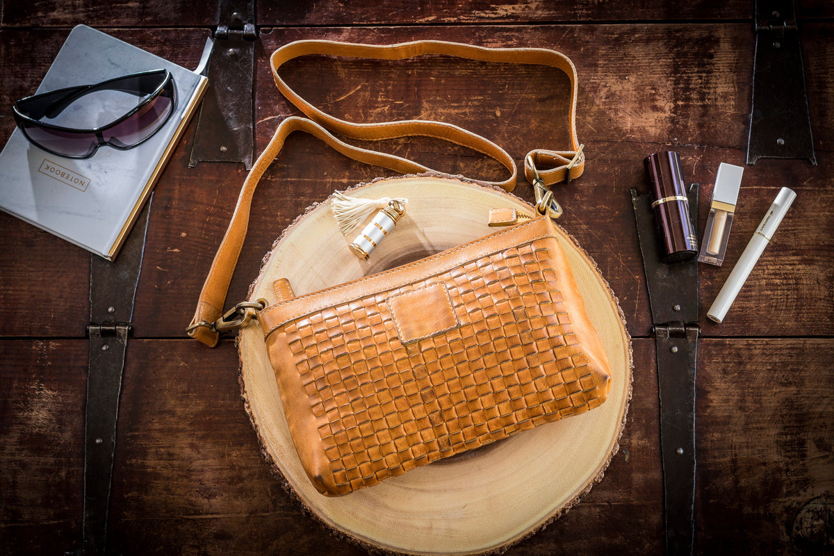 Tan woven crossbody bag on a wood round with its strap, sunglasses, a notebook and lipstick around it