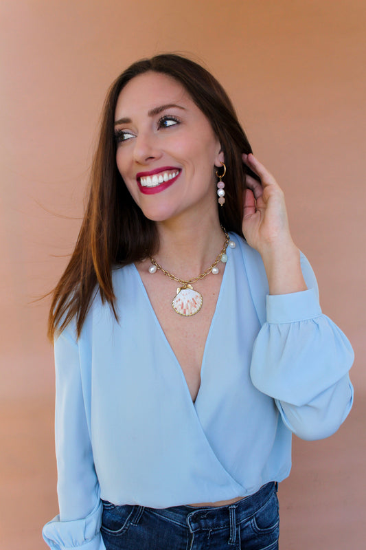 Smiling model in a pale blue wrap top wearing gemstone drop hoop earrings and a shell pendant