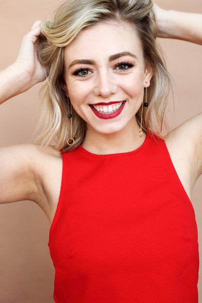 Smiling model in a red top, hands in her hair, wearing long chain drop earrings in a dark stone version