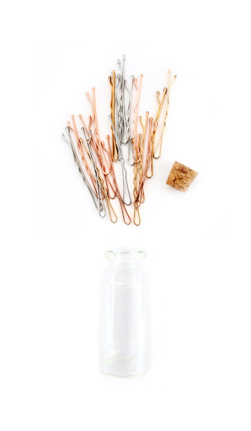 Mini bobby pins in gold tone, rose gold tone and silver tone spilling beside a glass canister and cork