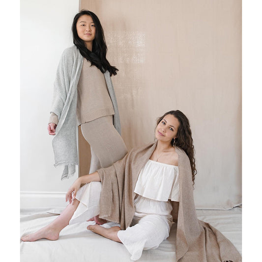Two models, one wearing the gray scarf open as a wrap and one seated under a taupe scarf