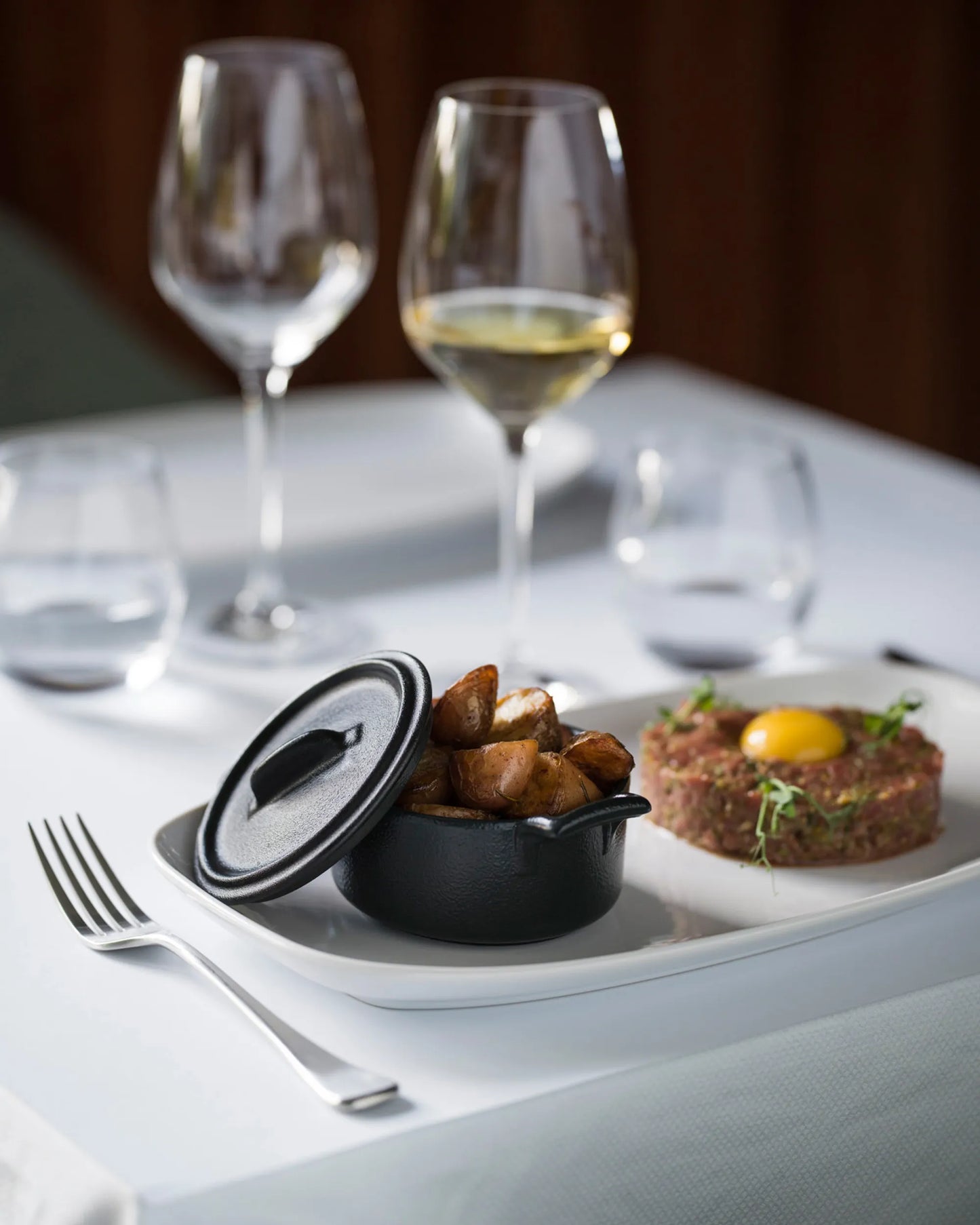 White linen table setting, a lidded black dish of potatoes beside a plated course.