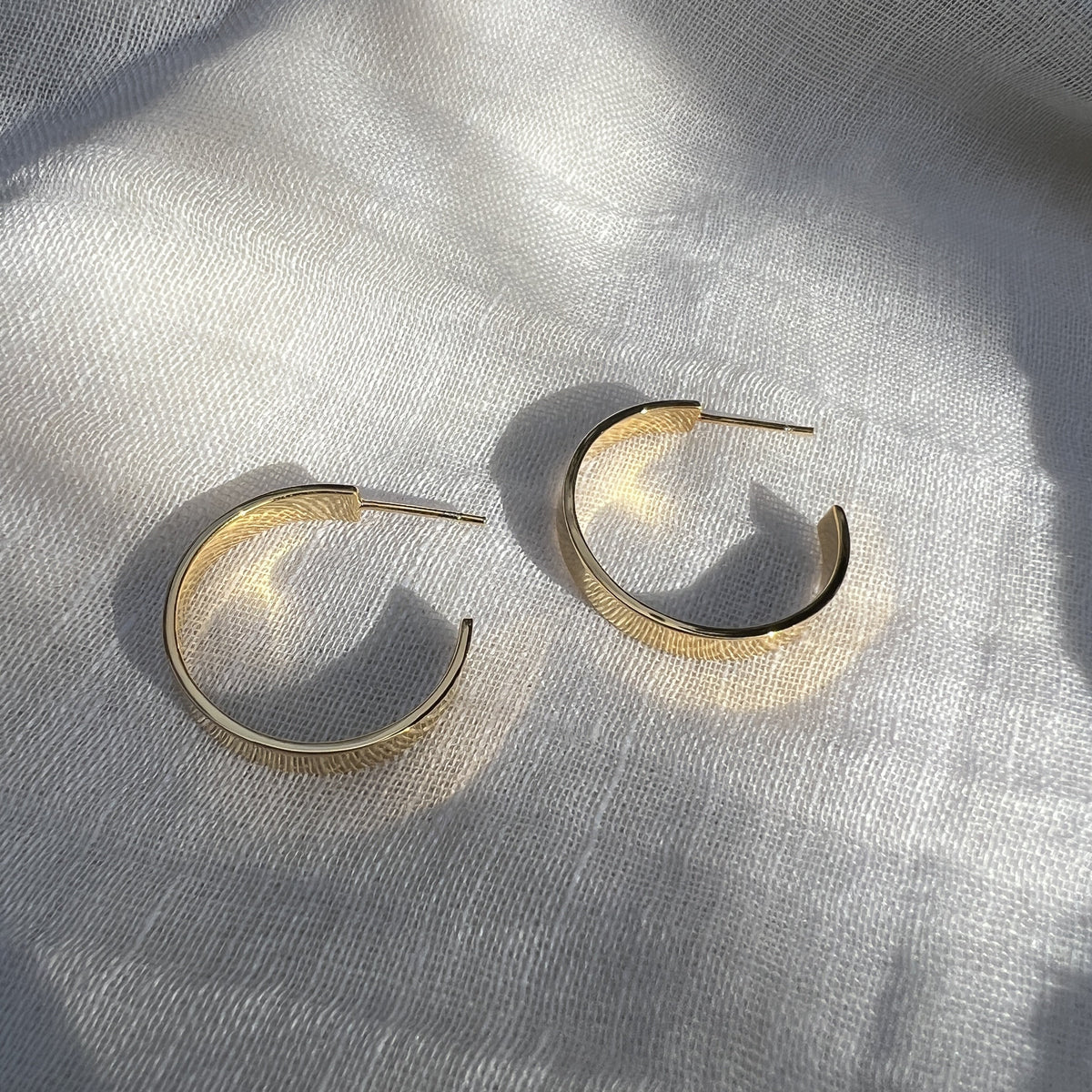 Chunky gold plated hoops resting on linen, the wide band catching the light along its edge
