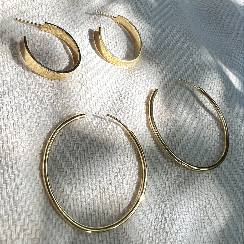 Chunky gold plated hoops beside a pair of thin large hoops on woven fabric in dappled light