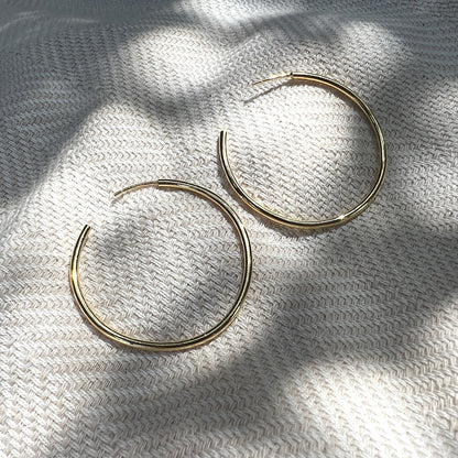 Pair of large gold plated hoop earrings on woven linen under leaf shadows