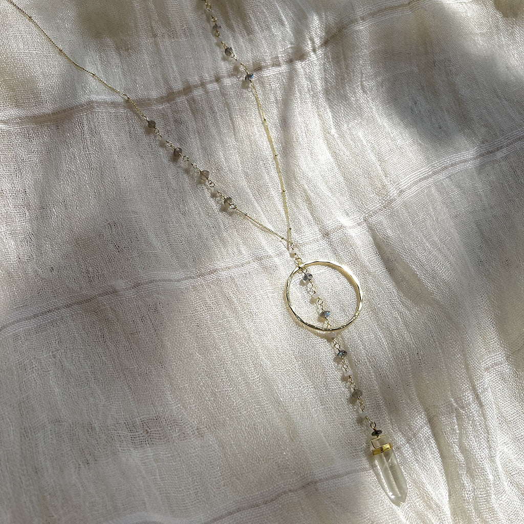 Clear quartz Y necklace laid on white gauze with soft shadows across the brass halo