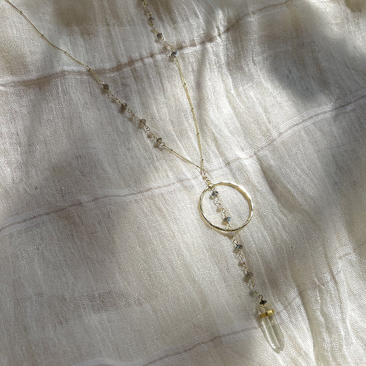 Clear quartz Y necklace laid on white gauze with soft shadows across the brass halo