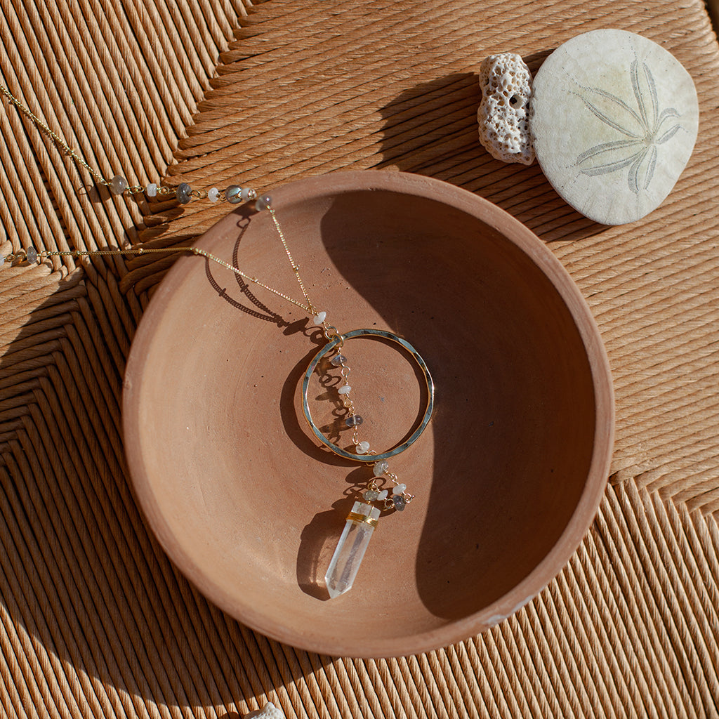 Clear quartz Y necklace in a terracotta bowl on a woven rush seat beside a sand dollar